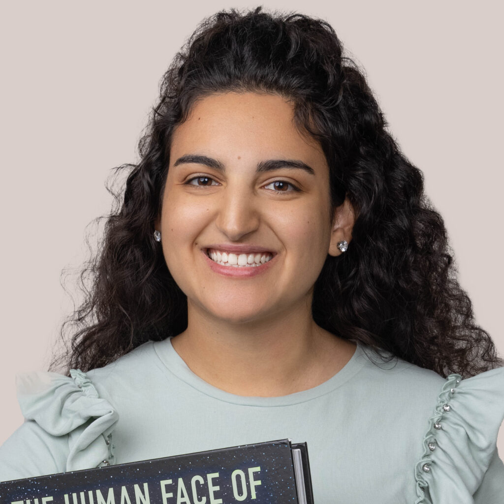 A young woman with curly dark hair and a bright, engaging smile. She is wearing a soft blue top with ruffle details on the sleeves and holds a book titled "The Human Face Of." The background is neutral, focusing attention on her cheerful expression and the book in her hands.