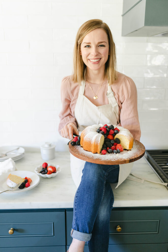 A woman (wearing a pink long-sleeve shirt, blue jeans, and a white apron) with short blonde hair sits on a gray countertop. She is holding a plate with an bundt cake that has mixed berries and powdered sugar on top.