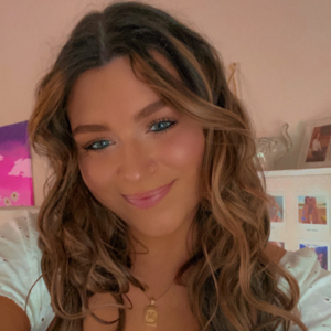 A smiling young woman with wavy brown hair and blue eyes, wearing a white blouse and a delicate necklace with a letter "M" pendant