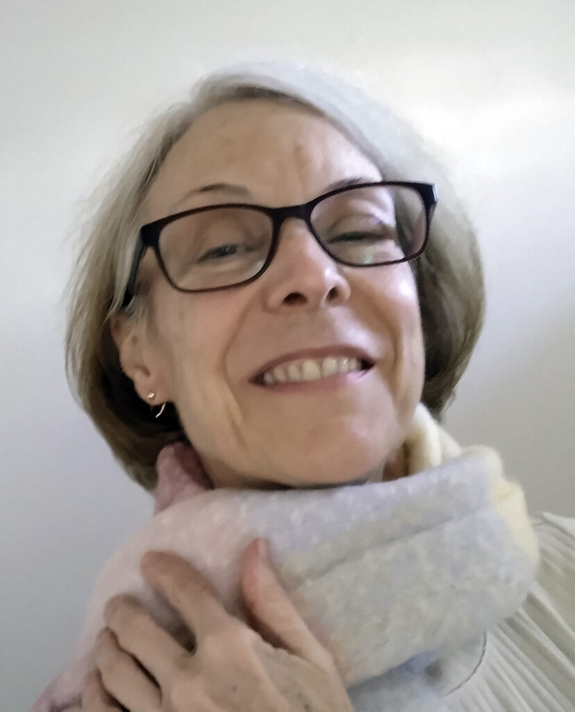 A cheerful woman with silver hair and black-framed glasses, smiling, wearing a pale scarf wrapped around her neck. The background is plain and bright.