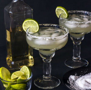 Two classic margarita cocktails with salted rims and lime wedges, served in stemmed glasses. A bottle of golden tequila and a small bowl with extra lime wedges and salt for garnish are set against a dark background, highlighting the clarity of the drinks.