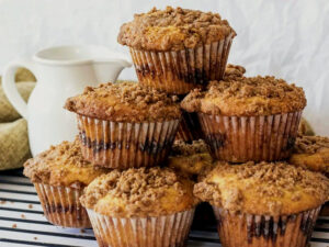 A stack of freshly baked muffins with a crumbly streusel topping, arranged to show their golden-brown texture. They're displayed on a wire rack with a white pitcher and a woven cloth in the background, suggesting a cozy, homemade setting.