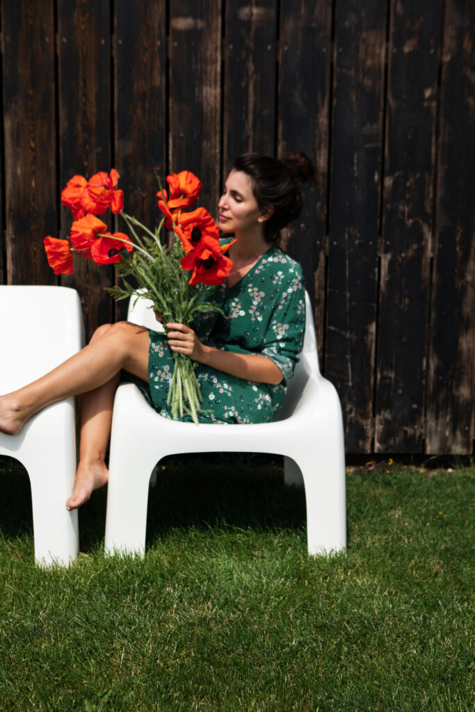 A woman in a green floral dress sitting on a modern white chair in a garden setting, holding a bunch of bright red poppies. She appears relaxed and contemplative, surrounded by lush green grass and a rustic wooden backdrop.