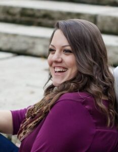 Portrait of a joyful woman sitting outdoors, dressed in a purple top, with a wide smile and curly brown hair. The background shows a blurred natural setting, enhancing the focus on her cheerful expression.