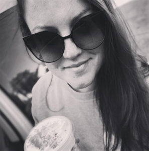 A woman in sunglasses holding a frosted drink, smiling subtly, and the image in black and white.