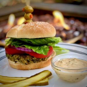 A veggie burger with a skewer of olives and peppers on top, served with a side of pickles and a dipping sauce on a white plate, set against a backdrop of a blazing fire.