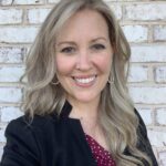 A woman with long, wavy blonde hair smiling in front of a light brick wall. She is wearing a black blazer over a red blouse with white polka dots, and gold hoop earrings. The setting is bright and casual, highlighting her friendly and approachable demeanor.