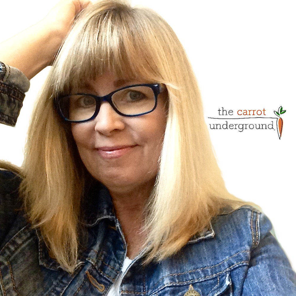 A smiling woman with blonde hair, wearing blue eyeglasses and a denim jacket, standing in front of a plain background with a logo that reads "the carrot underground".