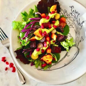 Colorful salad featuring beets, carrots, and pomegranate seeds drizzled with green avocado dressing on a decorative plate with leaf illustrations.