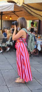 A woman with long hair wearing a bright, striped jumpsuit in red and pink is taking a photo with a DSLR camera.