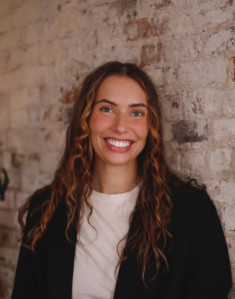 A portrait of a smiling woman with long, curly hair, standing against a textured brick wall. She wears a black blazer over a white top and has a bright, engaging smile, highlighting her friendly and approachable demeanor.