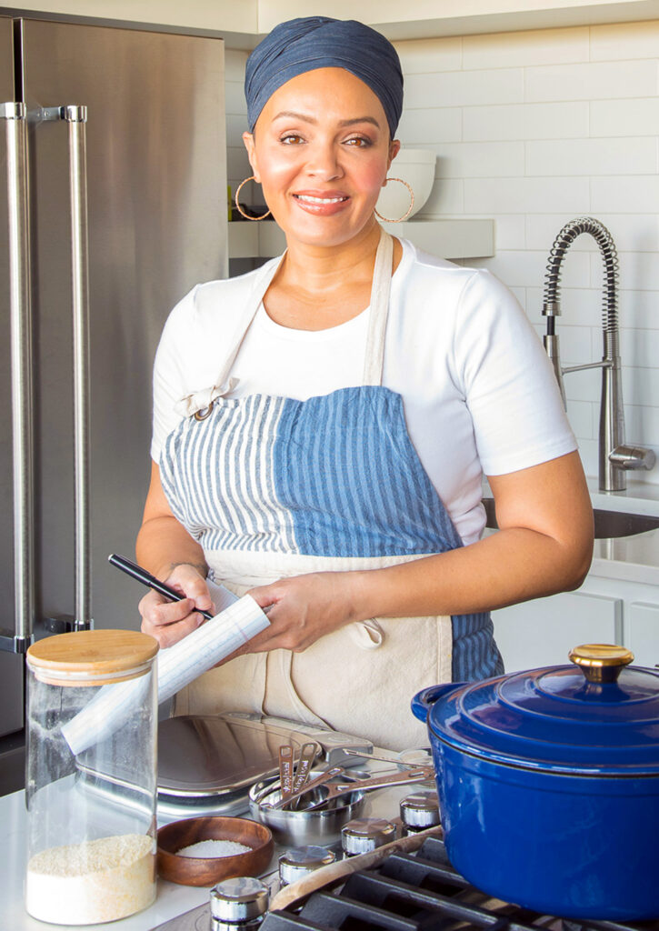 A professional photo of a smiling woman in a kitchen setting, wearing a blue headscarf and a blue and white striped apron. She is writing on a notebook, surrounded by kitchen utensils and a modern kitchen setup, indicating a culinary professional at work.
