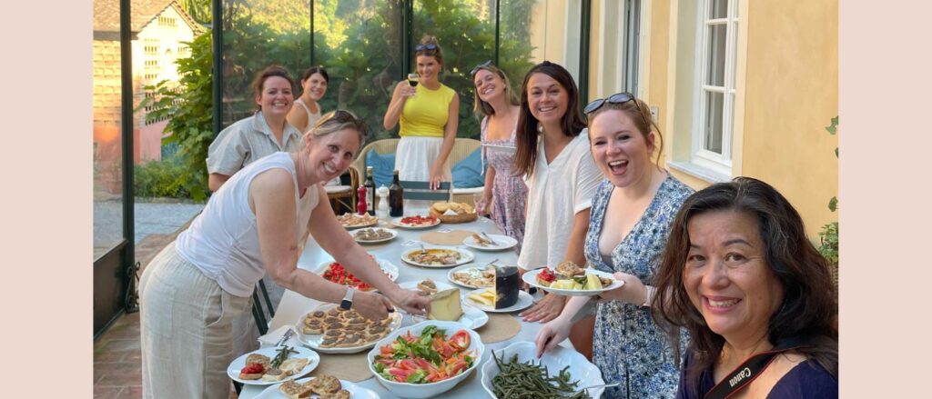 A group of smiling people gathered around a long table set with various dishes, including salads, bread, and other foods. The table is outdoors under a glass-covered patio, with greenery visible in the background