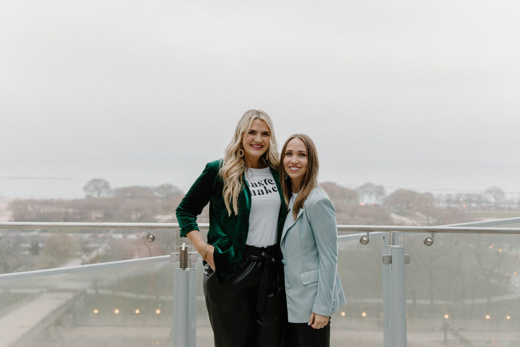 Two women standing together and smiling on an outdoor balcony with a foggy landscape in the background.