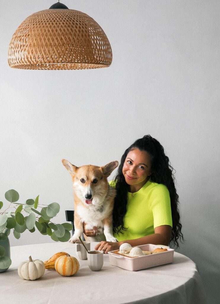 A woman with long, curly hair is sitting at a round table with a corgi dog beside her. She is wearing a bright neon green top and smiling gently at the camera. The table is decorated with small pumpkins and eucalyptus leaves, and a light-colored casserole dish is placed in front of her.