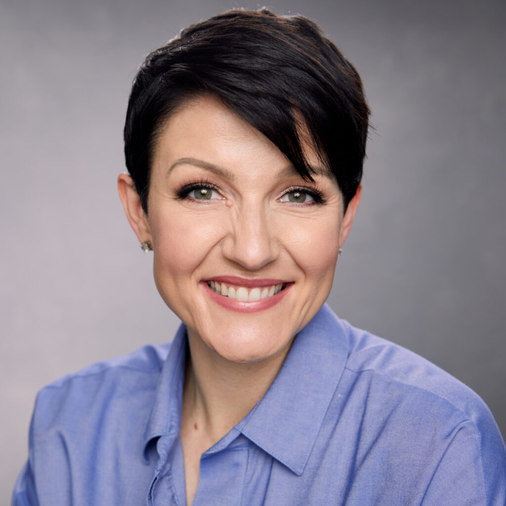 A woman with short, dark hair smiles warmly at the camera in a close-up headshot. She is wearing a light blue button-up shirt.