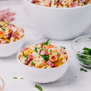 A colorful bowl of muffaletta pasta salad with diced meats, olives, and fresh herbs, served in a white bowl with a larger bowl of the same salad in the background.