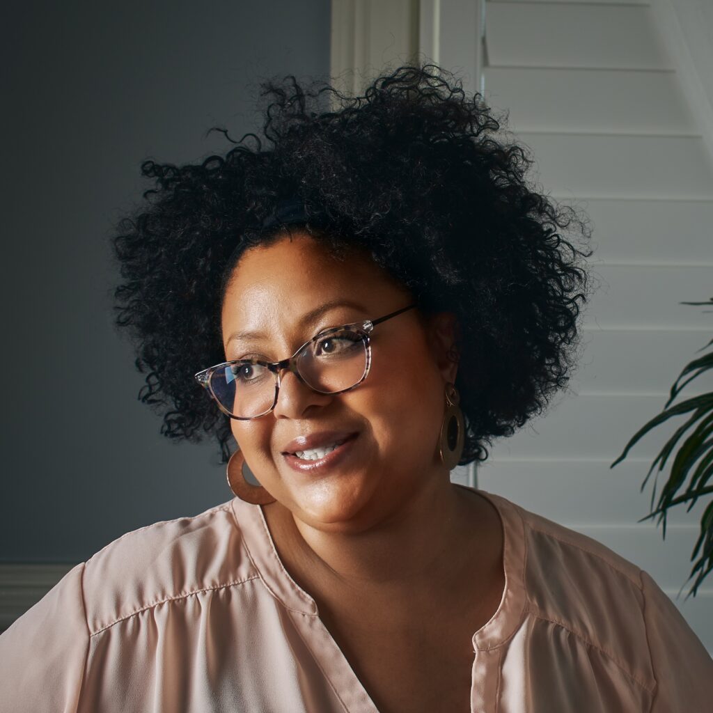 A portrait of a woman with curly black hair, wearing glasses and a light pink blouse, smiling softly in front of a neutral background with soft lighting.