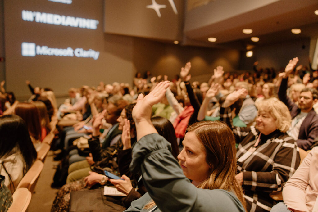 a large audience seated in a conference hall, with many people raising their hands