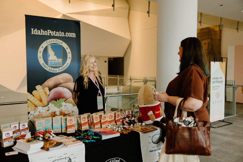 A smiling woman stands at an IdahoPotato.com booth, featuring the Certified 100% Idaho Potatoes logo, at a conference or trade show.