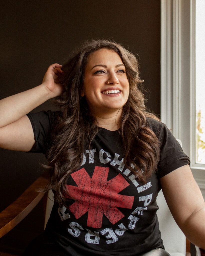 A candid portrait of a woman with long, wavy hair smiling and wearing a Red Hot Chili Peppers t-shirt, sitting near a window with soft lighting illuminating her face.