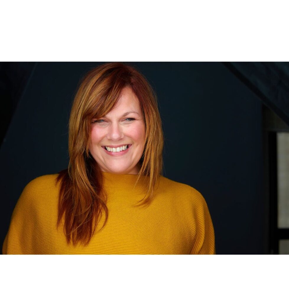 Portrait of a smiling woman with shoulder-length reddish-brown hair, wearing a mustard-yellow sweater. She has a bright, cheerful expression