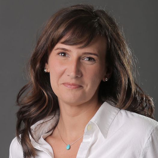 A professional headshot of Tara Long, featuring her wearing a white blouse and a turquoise necklace, smiling softly against a gray background with soft lighting.