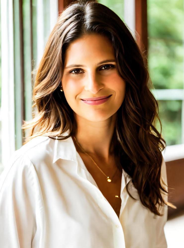 Portrait of a woman with medium-length brunette hair, wearing a white blouse. She is smiling gently and looking directly at the camera, standing in a room with a blurred green outdoor background visible through a window behind her.