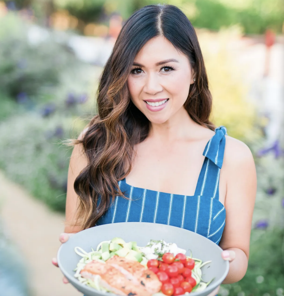 A woman with long, wavy dark brown hair smiles at the camera while holding a bowl of salad.