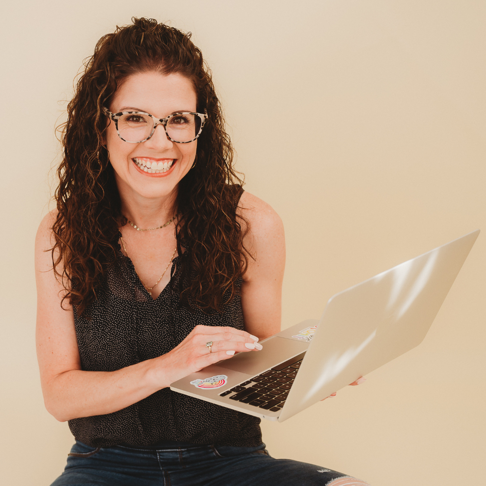 Portrait of Jennifer Holt, a woman with curly hair and glasses, smiling while holding a laptop with stickers. She is wearing a sleeveless top and sitting against a light-colored background.