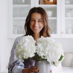 Liane Walker smiling in a bright kitchen while holding a vase filled with white hydrangeas.