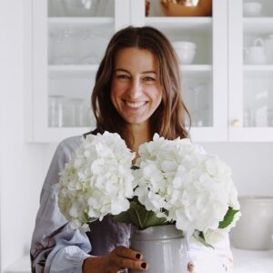 Liane Walker smiling in a bright kitchen while holding a vase filled with white hydrangeas.