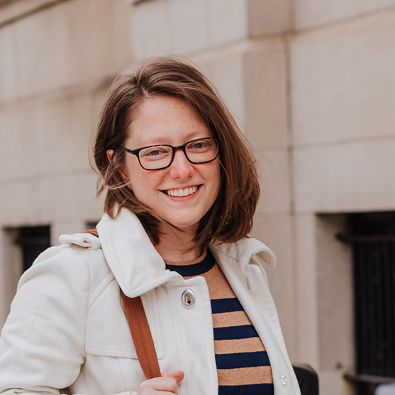 A smiling woman with glasses, wearing a white jacket, standing outdoors against a light stone building. She has shoulder-length brown hair and carries a brown bag.