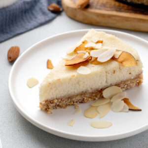 A slice of almond cheesecake on a white plate, topped with toasted almond slices, with scattered almonds and a wooden serving board in the background.