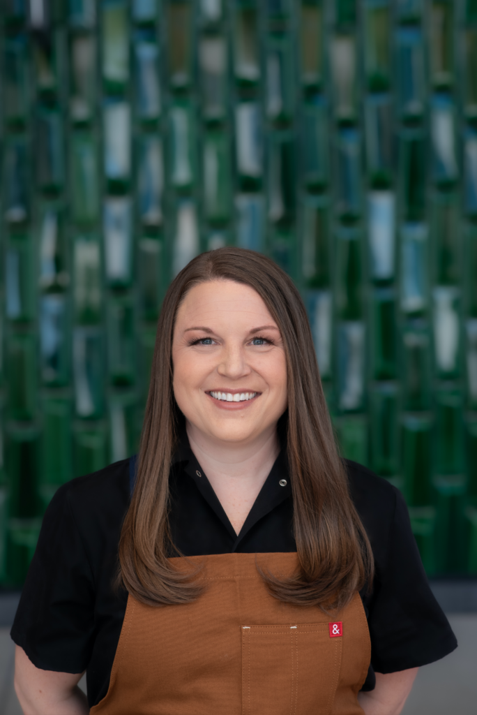 A woman with long brown hair smiling warmly while wearing a black shirt and a brown apron, standing in front of a backdrop of vertically aligned green bottles.