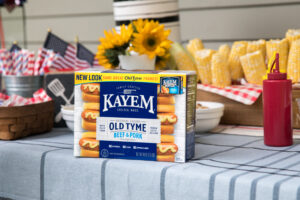A package of Kayem Old Tyme Beef & Pork Franks on a picnic table with a festive background, including sunflowers, American flags, corn on the cob, and a red ketchup bottle.