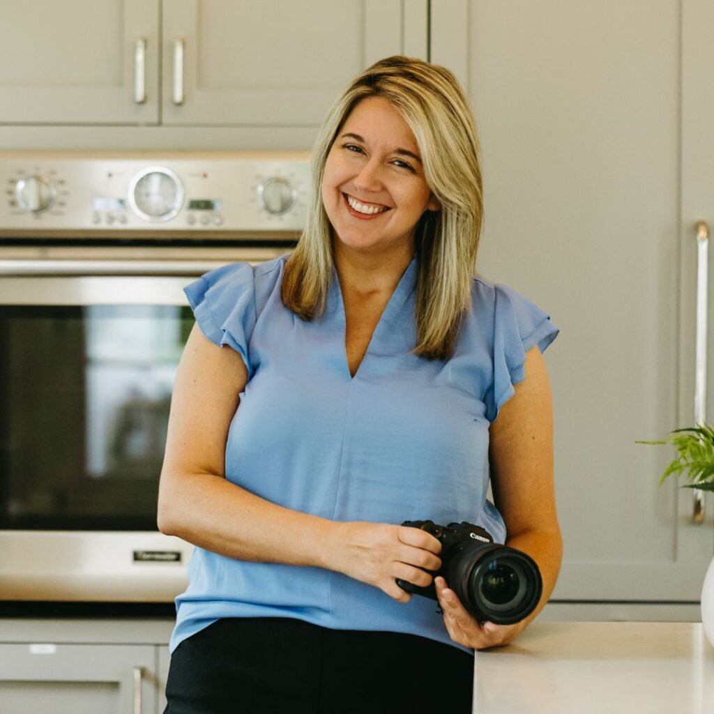 A woman with blonde hair smiling warmly while holding a camera in a modern kitchen, standing in front of a stainless steel oven and light gray cabinetry.