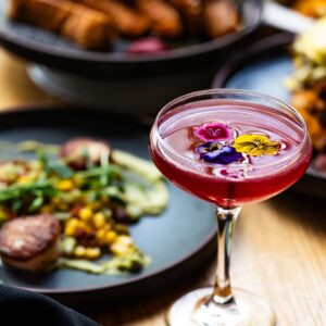 A vibrant purple cocktail in a coupe glass, garnished with colorful edible flowers, placed on a table with blurred gourmet dishes in the background.