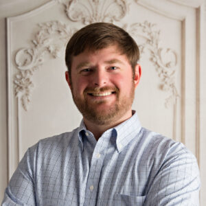 A professional headshot of a smiling man with short brown hair and a beard, wearing a light blue checkered shirt, standing in front of an ornate white background.