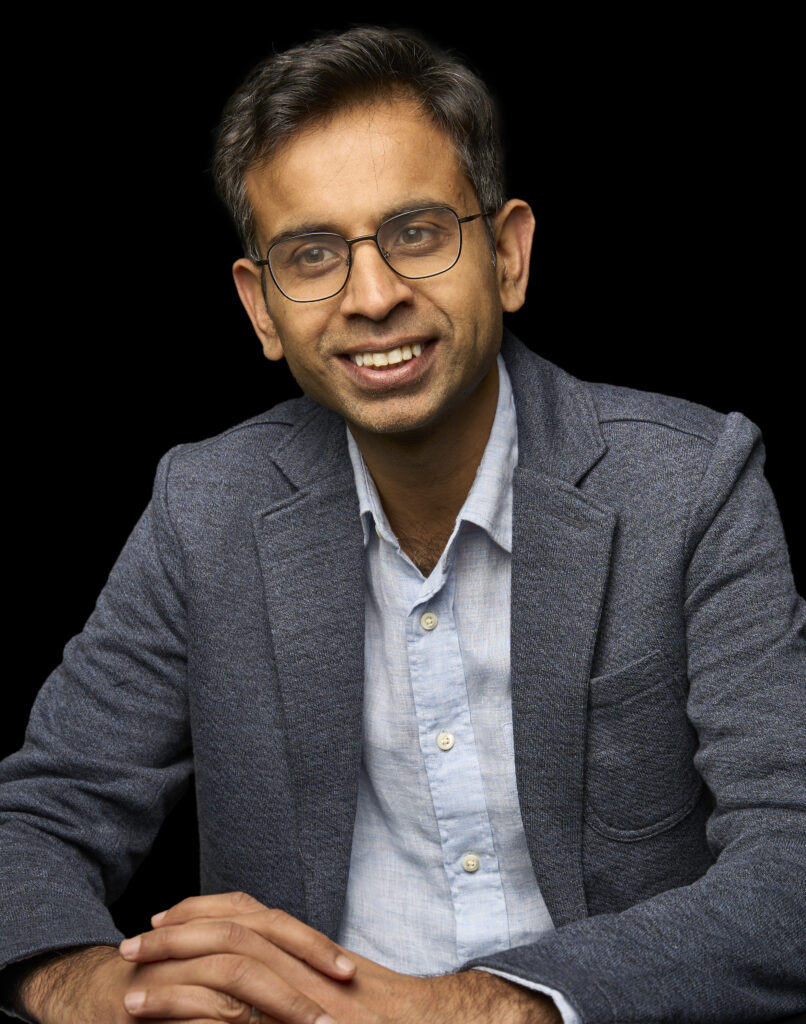 Portrait of a smiling man wearing glasses and a blue blazer over a light blue shirt, sitting with hands clasped on a black background.