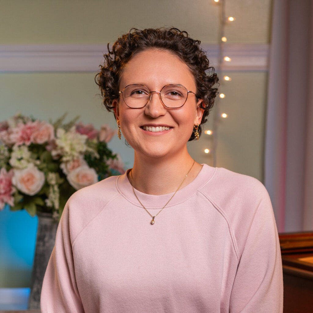 A photo of a woman with short curly brown hair and glasses, wearing a soft pink sweater. She is smiling warmly against a softly lit background featuring floral arrangements and string lights, creating a cozy and welcoming ambiance.