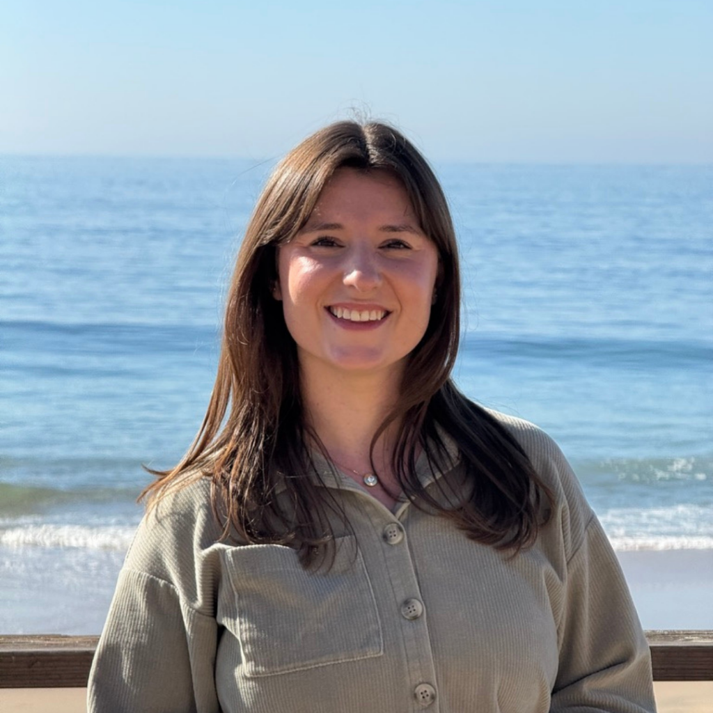 A woman with long brown hair smiles at the camera while standing in front of the ocean. She is wearing a sage green button-up shirt, and the sunlight highlights her face. The background features a clear blue sky and gentle ocean waves.