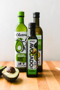 A trio of avocado oil bottles from different brands, including 'Chosen Foods' and 'AvoHass Organic Avocado Oil,' arranged on a wooden countertop. A halved avocado sits in the foreground, showcasing its seed and flesh.