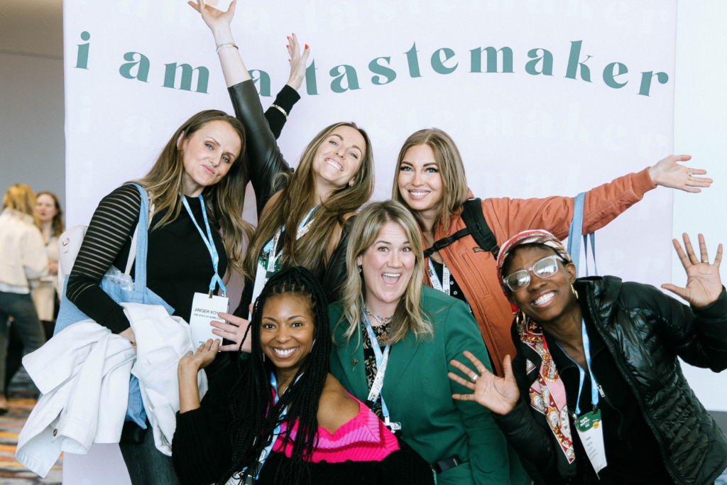 A group of six diverse women pose excitedly in front of a pastel-colored backdrop that reads 'I am a Tastemaker.'