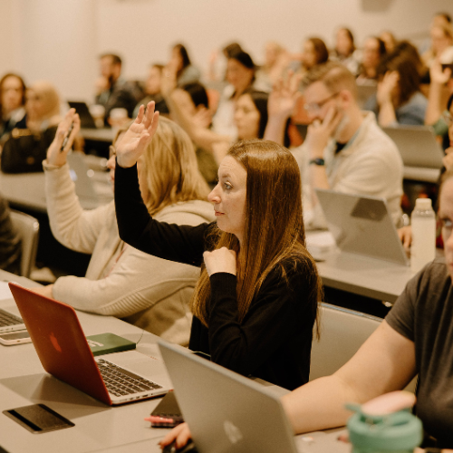 A classroom or conference session with attendees seated at long tables with laptops open. A woman in the foreground with long brown hair, wearing a black sweater, raises her hand while looking towards the front of the room.