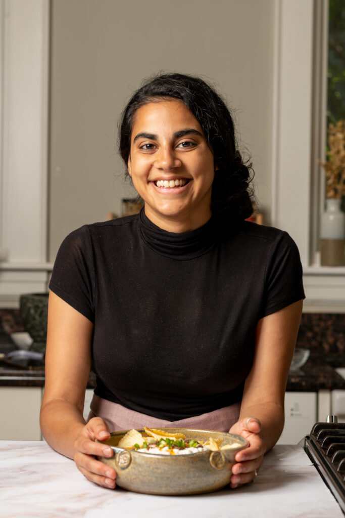 A smiling woman with dark hair tied back, wearing a black turtleneck, is holding a ceramic bowl filled with food. She is seated at a kitchen counter with a marble surface, with a stove and kitchen decor visible in the background.