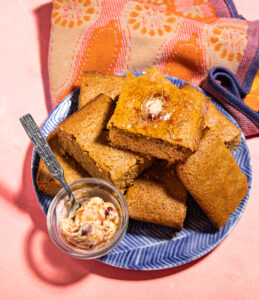 A plate of golden brown cornbread squares topped with melting butter and honey, accompanied by a small dish of whipped butter with visible fruit swirls, all set on a blue patterned plate with a colorful orange and pink textile in the background.