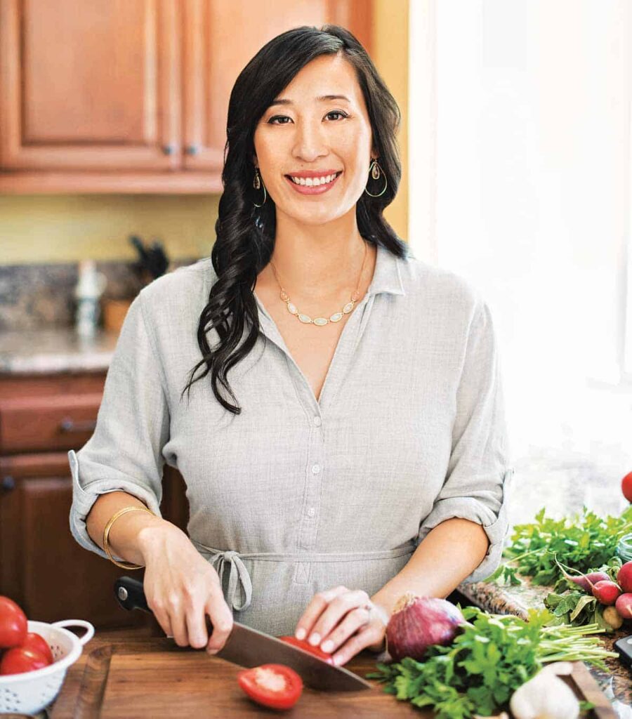 Jessica Gavin smiling in a bright kitchen while slicing a tomato on a wooden cutting board. She is wearing a light gray button-up dress with rolled sleeves, gold jewelry, and is surrounded by fresh vegetables including parsley, onions, and radishes."