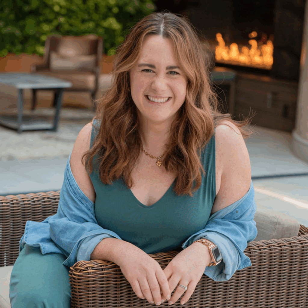 Bree Pair sitting outdoors in a wicker chair, smiling warmly at the camera. She is wearing a teal sleeveless dress with a light denim shirt draped over her shoulders, a gold necklace, and a smartwatch. A fire feature and greenery are visible in the background, creating a relaxed and inviting atmosphere.