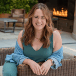 Bree Pair sitting outdoors in a wicker chair, smiling warmly at the camera. She is wearing a teal sleeveless dress with a light denim shirt draped over her shoulders, a gold necklace, and a smartwatch. A fire feature and greenery are visible in the background, creating a relaxed and inviting atmosphere.
